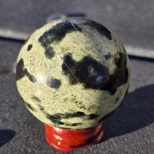 May include: A speckled green and black stone sphere, resting on a small, glossy red base. The sphere has a mottled pattern of dark green and black spots against a lighter green background. The base is a vibrant red.