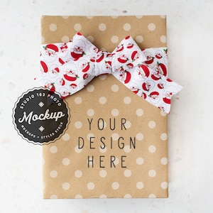 May include: A wrapped gift with a brown paper cover and white polka dots. A decorative bow with a Santa Claus pattern in red, white, and black is tied around the package. The text "YOUR DESIGN HERE" is printed on the front.