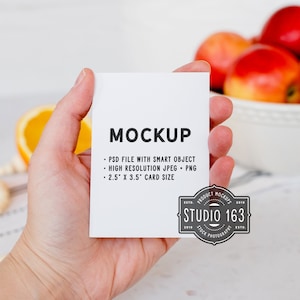 May include: A white card mockup with the text "MOCKUP" and details about the file type and size. The card is being held by a hand against a white background with a bowl of fruit and a string of beads in the background.