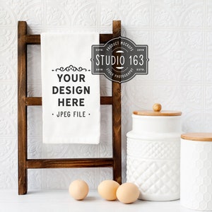 May include: A white kitchen towel with the text "YOUR DESIGN HERE" and "JPEG FILE" hanging on a wooden ladder. There are two white ceramic canisters with wooden lids and three brown eggs on a white surface.