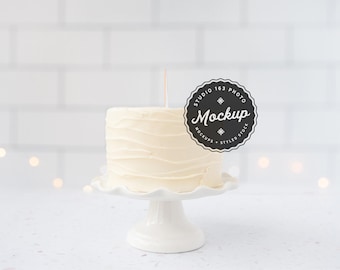 May include: A white cake with a simple buttercream frosting sits on a white cake stand. A black and white circle sticker with the text "Studio 163 Photo Mockup" is attached to the cake.