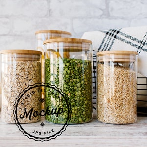 May include: Four glass jars with bamboo lids filled with various dry goods, including oats, green peas, and rice. The jars are sitting on a white wooden surface with a white and black checked cloth and a black wire basket in the background.