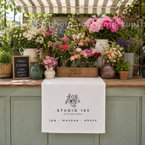 May include: A flower stand displays a variety of fresh cut flowers in vases and containers. A chalkboard sign reads "FRESH FLOWERS, STEMS, BOUQUETS, PLANTS." A wooden box reads "FRESH CUT FLOWERS." A white tablecloth features a floral design and the text "STUDIO 163 photography jpg mockup photo."