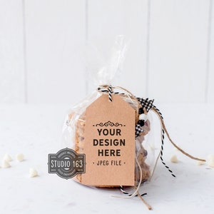 May include: A clear plastic bag filled with cookies tied with twine and a brown gift tag. The tag has the text "YOUR DESIGN HERE . JPEG FILE." printed on it. White chocolate chips are scattered on a white surface.