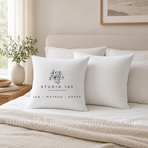 Op de afbeelding: Twee witte vierkante kussens op een bed met een beige sprei. Eén kussen heeft een bloemenontwerp en de tekst "STUDIO 163 photography jpg mock up photo". Het bed is opgemaakt met witte lakens.