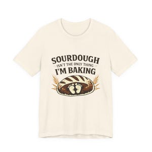 May include: Cream-colored t-shirt with the text "SOURDOUGH ISN'T THE ONLY THING I'M BAKING" above a graphic of a loaf of bread with baby footprints. Two wheat stalks flank the bread graphic.