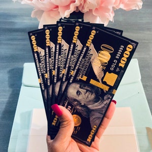 Puede incluir: Una mano sosteniendo una pila de billetes de 1000 € negros y dorados con el texto "GOLD 999999" en el billete superior.