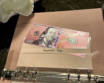 May include: A pink binder with a clear plastic pocket containing a fake $500 bill with the text "Household/Preps" written on the pocket.