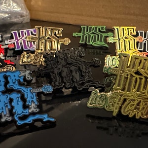 May include: A collection of nine enamel pins featuring the text "Lost Cause" in various colors, including red, purple, gold, green, black, and blue. The pins are designed with a dripping effect and feature a syringe and guitar.