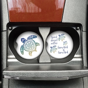 May include: Two round car coasters with a white background. One coaster features a colorful mandala-style sea turtle design. The other coaster features a colorful sea turtle design with the text "Speed doesn't matter forward is forward".