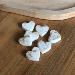 May include: Six white heart-shaped wax melts with gold glitter. The melts are on a wooden surface.