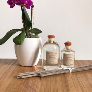 Könnte beinhalten: Zwei klare Glasflaschen mit braunen Kappen und Etiketten mit der Aufschrift "Diffuseur de parfum Rose Des Jardins Fabrication artisanale". Die Flaschen stehen auf einer Holzoberfläche mit einem Bündel grauer Stäbchen. Im Hintergrund befindet sich eine Orchidee im Topf mit rosa Blüten.