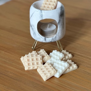 Op de afbeelding: Een witte marmeren wax warmer met gouden poten en een witte theelichtje. Er zijn zes wax melts in de vorm van wafels, drie zijn wit en drie zijn beige.