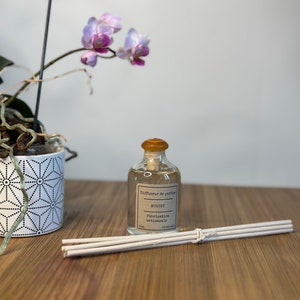 以下が含まれることがあります： 透明なガラス瓶に、茶色のラベルが貼ってあり、「Diffuseur de parfum MUGUET Fabrication artisanale 50ml」と書いてあります。瓶は木製の表面に置かれ、その横に白い棒が置かれています。背景には、ピンクの花を咲かせた鉢植えの蘭があります。