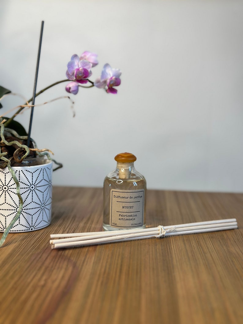 以下が含まれることがあります： 透明なガラス瓶に、茶色のラベルが貼ってあり、「Diffuseur de parfum MUGUET Fabrication artisanale 50ml」と書いてあります。瓶は木製の表面に置かれ、その横に白い棒が置かれています。背景には、ピンクの花を咲かせた鉢植えの蘭があります。
