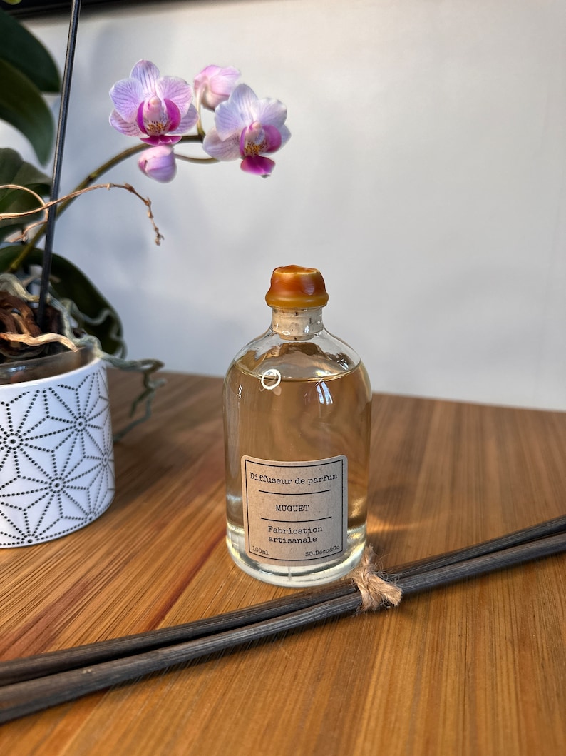 以下が含まれることがあります： 茶色のコルク栓と茶色のラベルが付いた透明なガラス瓶。ラベルには「Diffuseur de parfum, Muguet, Fabrication artisanale, 100ml, So.Decoco.」と書いてあります。