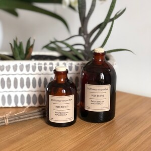 May include: Two brown glass diffuser bottles with cork stoppers. Each bottle has a white label with black text that reads "BOIS DE OUD" and "Fabrication artisanale". The bottles are on a wooden surface, with plants in the background.