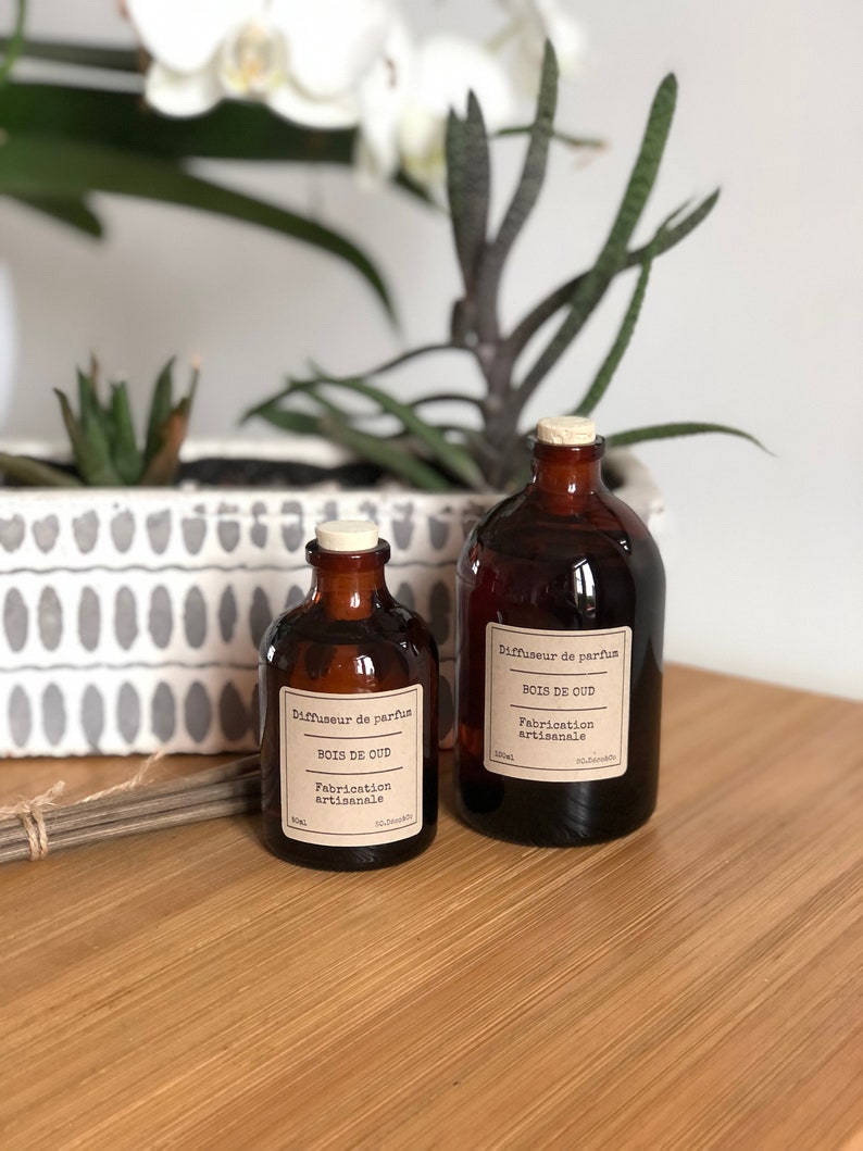 May include: Two brown glass diffuser bottles with cork stoppers. Each bottle has a white label with black text that reads "BOIS DE OUD" and "Fabrication artisanale". The bottles are on a wooden surface, with plants in the background.