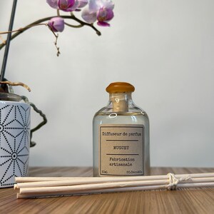 以下が含まれることがあります： 茶色のコルク栓と茶色のラベルが付いた透明なガラス瓶。ラベルには「Diffuseur de parfum MUGUET Fabrication artisanale 50mL 50.Deco&Co」と書いてあります。瓶は木製の表面に置かれ、木製の棒の束が置かれています。