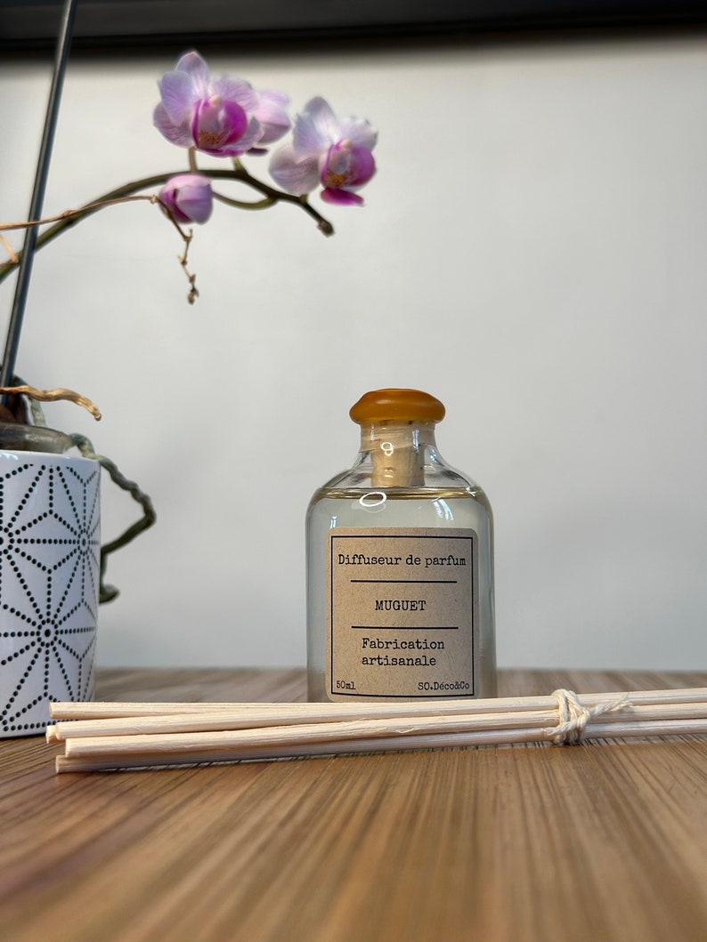 以下が含まれることがあります： 茶色のコルク栓と茶色のラベルが付いた透明なガラス瓶。ラベルには「Diffuseur de parfum MUGUET Fabrication artisanale 50mL 50.Deco&Co」と書いてあります。瓶は木製の表面に置かれ、木製の棒の束が置かれています。