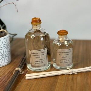 以下が含まれることがあります： ブラウンの蓋と「Diffuseur de parfum」と「Muguet」と書かれたラベルが付いたガラス瓶が2本。瓶は透明な液体で満たされ、木製の表面の上に置かれています。小さい方の瓶は50ml、大きい方の瓶は100mlです。瓶の前に木製のスティックがあります。