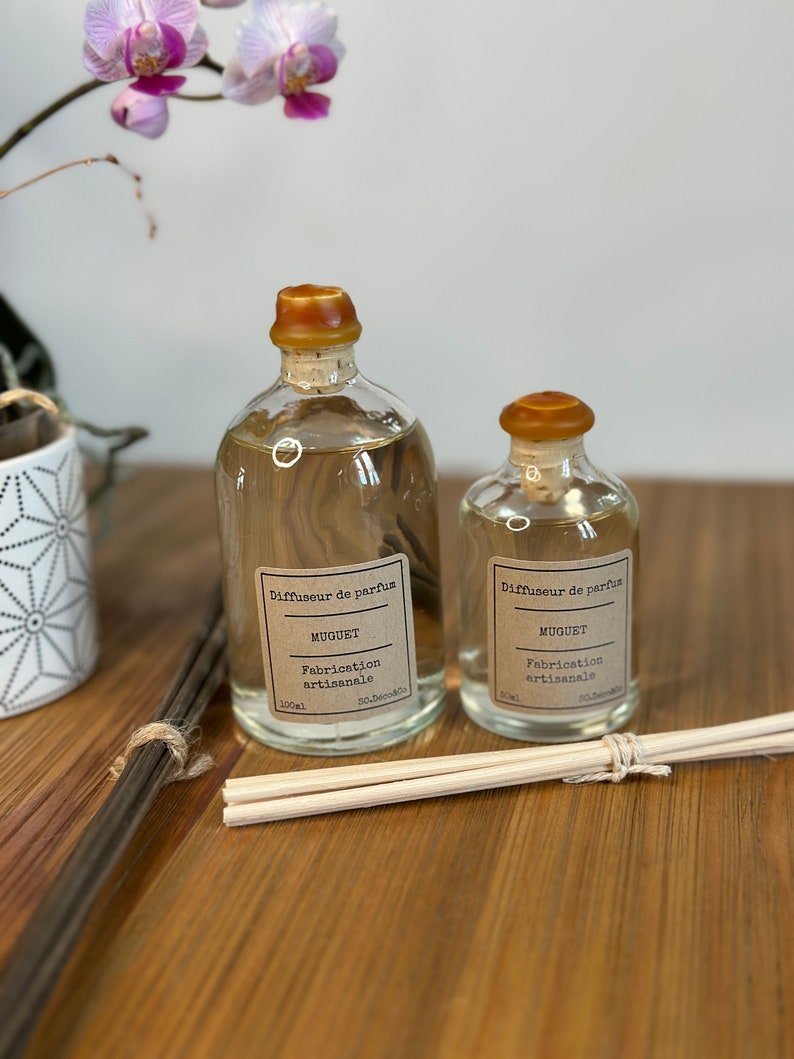 以下が含まれることがあります： ブラウンの蓋と「Diffuseur de parfum」と「Muguet」と書かれたラベルが付いたガラス瓶が2本。瓶は透明な液体で満たされ、木製の表面の上に置かれています。小さい方の瓶は50ml、大きい方の瓶は100mlです。瓶の前に木製のスティックがあります。