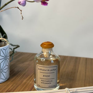 以下が含まれることがあります： 茶色のラベルに「Diffuseur de parfum MUGUET Fabrication artisanale 50 ml So.DecoSo.」と書いてある透明なガラス瓶。コルク栓が付いており、木製の棒束が置かれた木製の表面の上に置かれている。