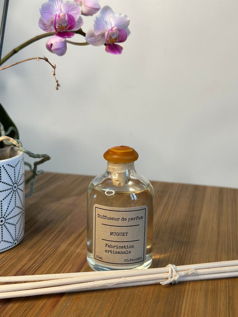 以下が含まれることがあります： 茶色のラベルに「Diffuseur de parfum MUGUET Fabrication artisanale 50 ml So.DecoSo.」と書いてある透明なガラス瓶。コルク栓が付いており、木製の棒束が置かれた木製の表面の上に置かれている。