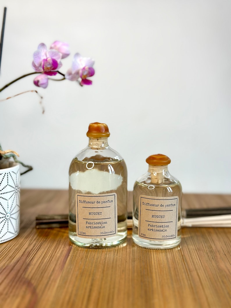 以下が含まれることがあります： コルク栓と茶色の紙のラベルが付いたガラス瓶が2本。ラベルには「Diffuseur de parfum」と「MUGUET」と書かれています。瓶は透明な液体で満たされており、木製の表面の上に置かれています。