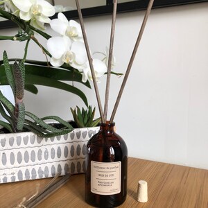 May include: A brown glass diffuser bottle with reeds, labeled "Bois de Oud" and "Fabrication artisanale." A white stopper sits beside the bottle. The background includes white orchids and a decorative planter with succulents.