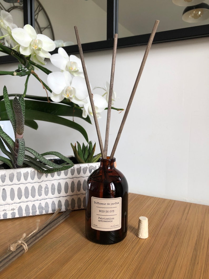 May include: A brown glass diffuser bottle with reeds, labeled "Bois de Oud" and "Fabrication artisanale." A white stopper sits beside the bottle. The background includes white orchids and a decorative planter with succulents.