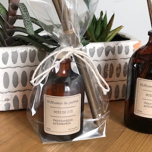 May include: A brown glass diffuser bottle with a label reading "BOIS DE OUD" and "Fabrication artisanale." The bottle is wrapped in clear cellophane and tied with twine, accompanied by wooden reeds. The product is 50ml.
