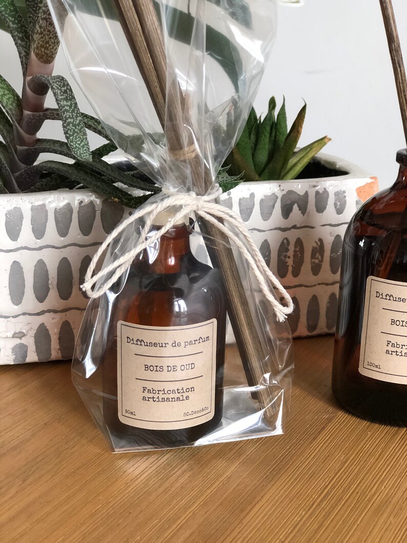 May include: A brown glass diffuser bottle with a label reading "BOIS DE OUD" and "Fabrication artisanale." The bottle is wrapped in clear cellophane and tied with twine, accompanied by wooden reeds. The product is 50ml.