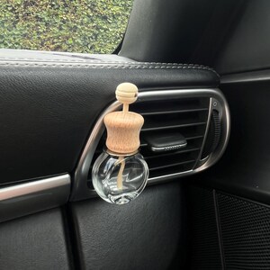 Peut inclure: Un diffuseur d'air de voiture en verre transparent avec un dessus en bois et un bâtonnet en bois à l'intérieur. Le diffuseur est suspendu à une bouche d'aération de voiture.
