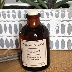 May include: A brown glass perfume diffuser bottle with a cork stopper. The label reads "BOIS DE OUD" and "Fabrication artisanale". The bottle is 50ml. The background includes a white planter with green plants.