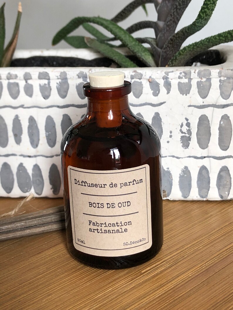 May include: A brown glass perfume diffuser bottle with a cork stopper. The label reads "BOIS DE OUD" and "Fabrication artisanale". The bottle is 50ml. The background includes a white planter with green plants.