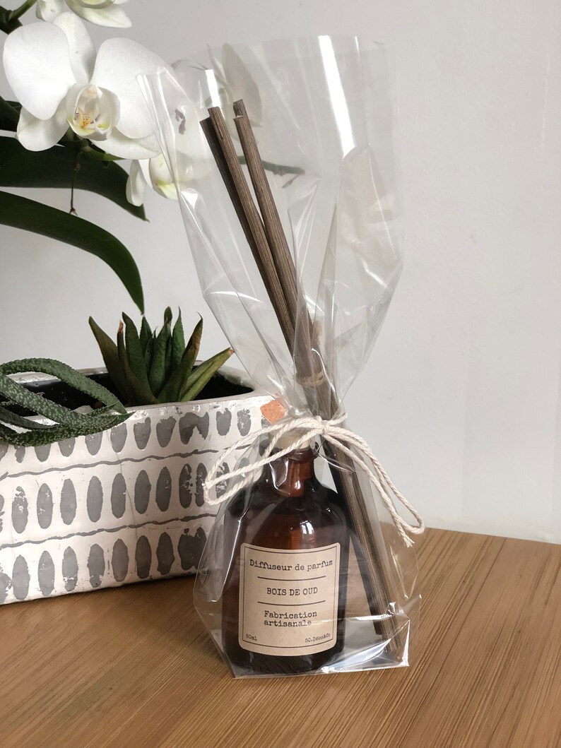 May include: A brown glass diffuser bottle with a label reading "Bois de Oud" and "Fabrication artisanale" is wrapped in clear cellophane and tied with twine. The bottle contains wooden reeds for fragrance diffusion. A white orchid and succulent plant are in the background.