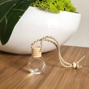 K&ouml;nnte beinhalten: Eine kleine, klare Glasflasche mit einem Holzverschluss und einer Schnur. Die Flasche h&auml;ngt an einem wei&szlig;en Keramiktopf mit gr&uuml;nem Moos.