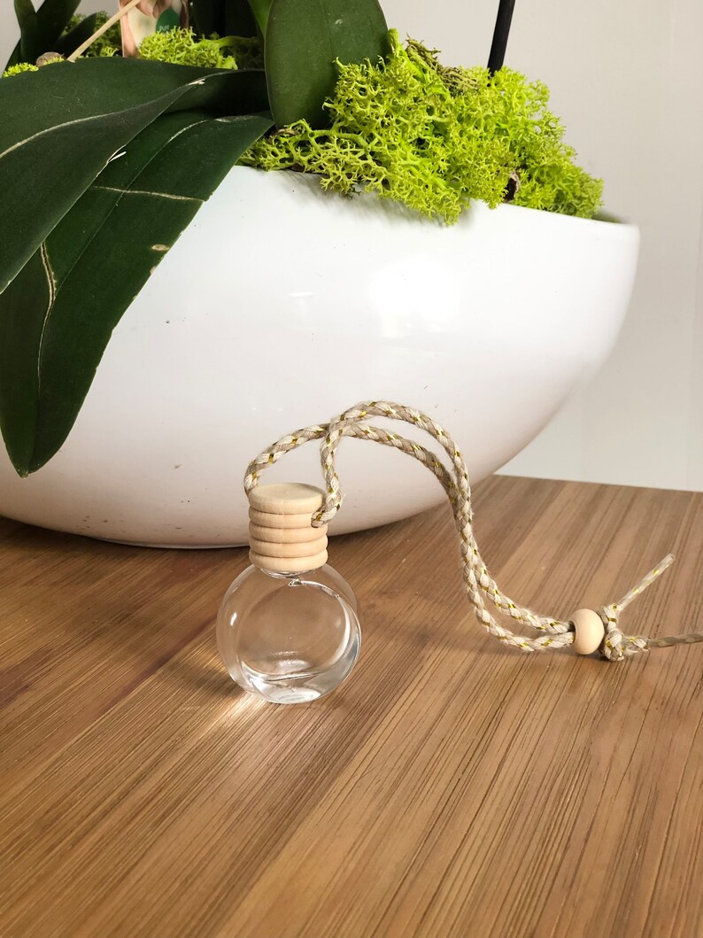 K&ouml;nnte beinhalten: Eine kleine, klare Glasflasche mit einem Holzverschluss und einer Schnur. Die Flasche h&auml;ngt an einem wei&szlig;en Keramiktopf mit gr&uuml;nem Moos.