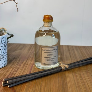 以下が含まれることがあります： 茶色のコルク栓と茶色のラベルが付いた透明なガラス瓶。ラベルには「Diffuseur de parfum MUGUET Fabrication artisanale 100ml SOL.DECORADO」と書いてあります。瓶は透明な液体で満たされており、黒い棒が置かれた木製の表面の上に置かれています。