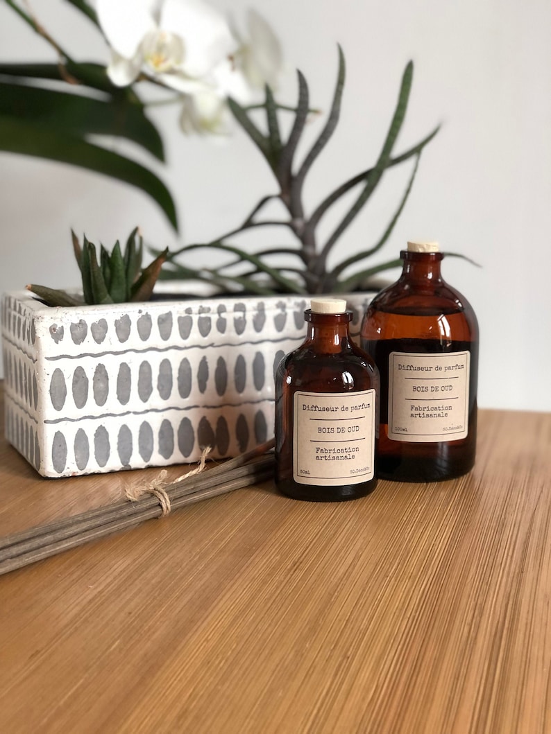 May include: Two amber glass diffuser bottles with cork stoppers and labels that read "BOIS DE OUD". A bundle of diffuser sticks and a white and gray patterned planter with greenery are also visible.