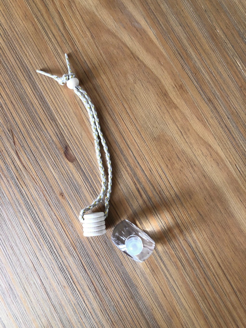 K&ouml;nnte beinhalten: Ein wei&szlig;es Band mit einer Holzperle oben und einem Stapel Holzringe unten. Neben dem Band befindet sich eine durchsichtige Glasflasche mit einem wei&szlig;en Verschluss.