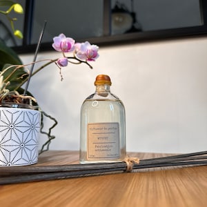 以下が含まれることがあります： 木製の表面に置かれた、茶色のラベルが付いた透明なガラス瓶。ラベルには「Diffuseur de parfum MUGUET Fabrication artisanale 100ml」と書いてあります。背景には黒い棒と鉢植えの蘭があります。