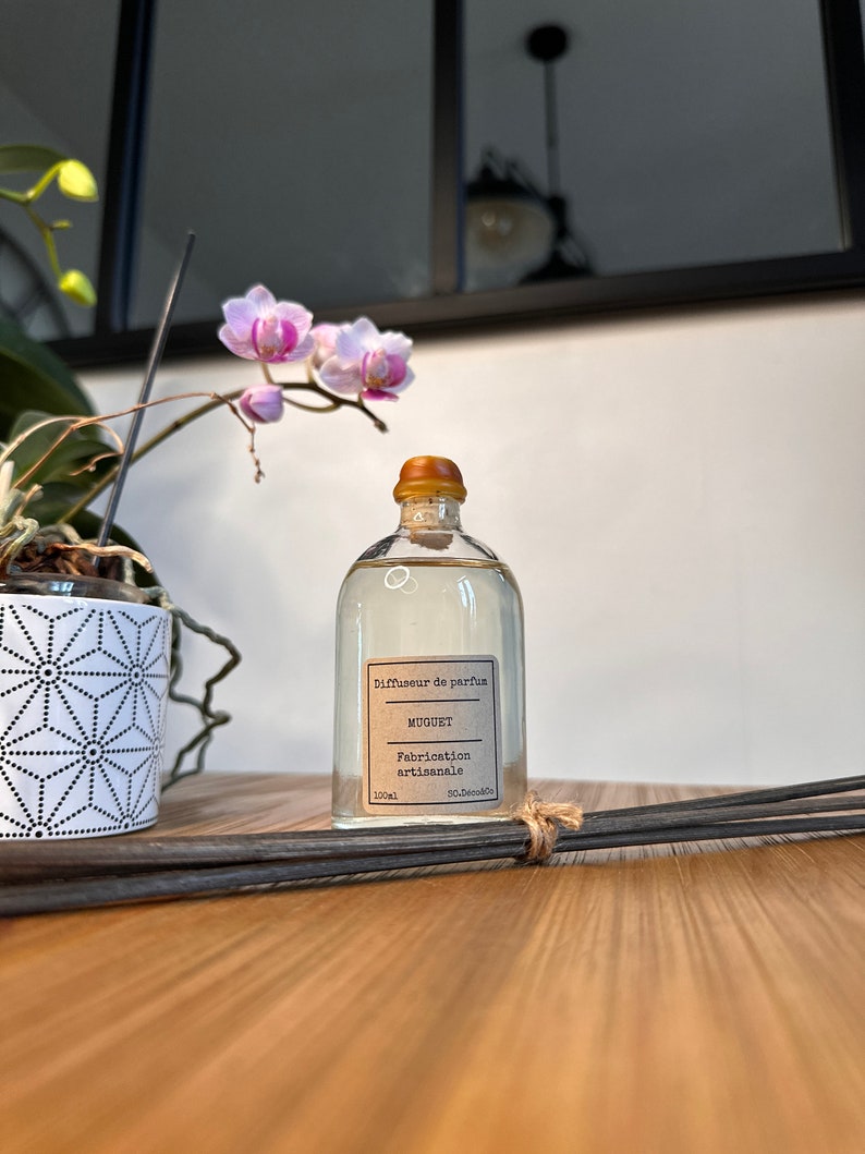 以下が含まれることがあります： 木製の表面に置かれた、茶色のラベルが付いた透明なガラス瓶。ラベルには「Diffuseur de parfum MUGUET Fabrication artisanale 100ml」と書いてあります。背景には黒い棒と鉢植えの蘭があります。