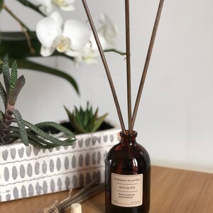 May include: A brown glass diffuser bottle with wooden reeds. The label reads "Bois de Oud, Fabrication artisanale." A bundle of reeds and a cork stopper are next to the bottle. White orchids and succulents are in the background.