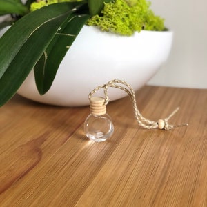 K&ouml;nnte beinhalten: Eine kleine, klare Glasflasche mit einem Holzstopfen und einer braunen Schnur, die daran h&auml;ngt. Die Flasche h&auml;ngt an einer wei&szlig;en Oberfl&auml;che.