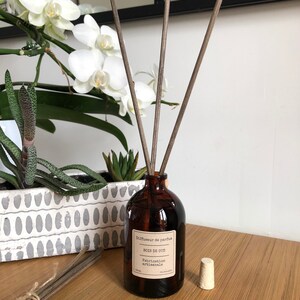 May include: A brown glass diffuser bottle with wooden reeds, a cork, and a label that reads "Bois de Oud". The bottle is on a wooden surface, with a bundle of reeds and a decorative planter with white flowers in the background.