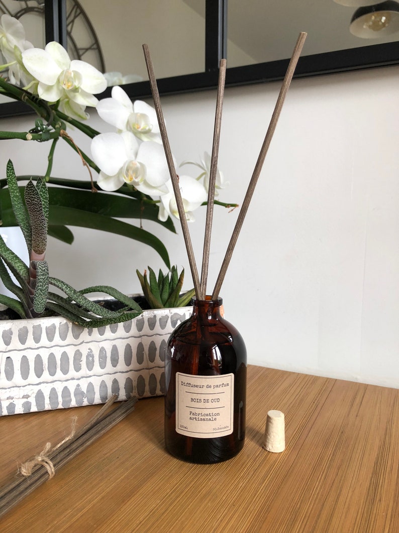 May include: A brown glass diffuser bottle with wooden reeds, a cork, and a label that reads "Bois de Oud". The bottle is on a wooden surface, with a bundle of reeds and a decorative planter with white flowers in the background.