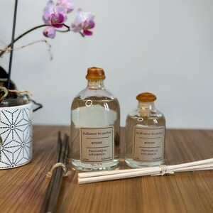 以下が含まれることがあります： フランス語で「Diffuseur de parfum, Muguet, Fabrication artisanale」と書かれた茶色の蓋とラベルが付いた透明なガラス瓶が2本。瓶は透明な液体で満たされ、茶色と白の棒の束が置かれた木製の表面の上に置かれています。