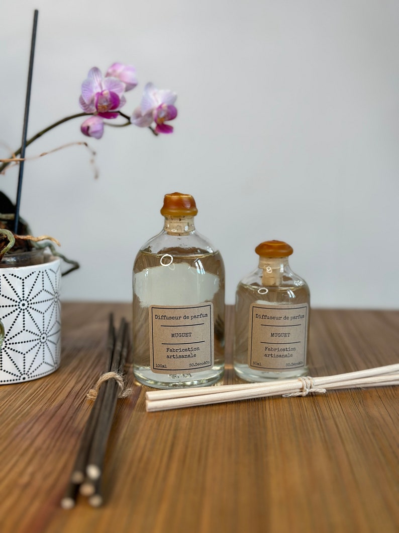 以下が含まれることがあります： フランス語で「Diffuseur de parfum, Muguet, Fabrication artisanale」と書かれた茶色の蓋とラベルが付いた透明なガラス瓶が2本。瓶は透明な液体で満たされ、茶色と白の棒の束が置かれた木製の表面の上に置かれています。